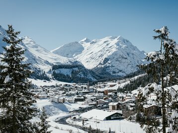 Winterliche Aussicht auf Lech am Arlberg, ein verschneites Bergdorf umgeben von majestätischen Alpen. Im Vordergrund ragen verschneite Nadelbäume in das Bild, während sich im Tal die Siedlung mit ihren schneebedeckten Dächern erstreckt. Im Hintergrund erheben sich schneebedeckte Berghänge und Gipfel unter einem klaren blauen Himmel. | © Ski Arlberg/Frederick Sams